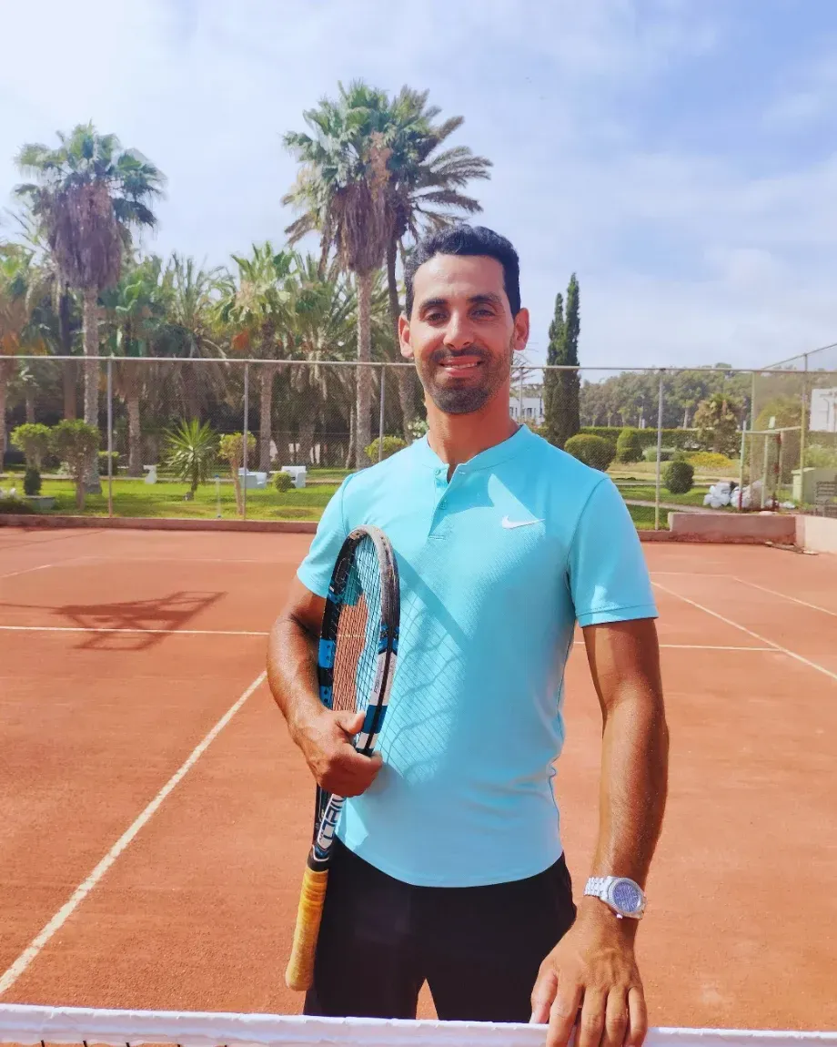 Le coach A.Mohamed, raquette à la main, au filet sur un court en terre battue du club