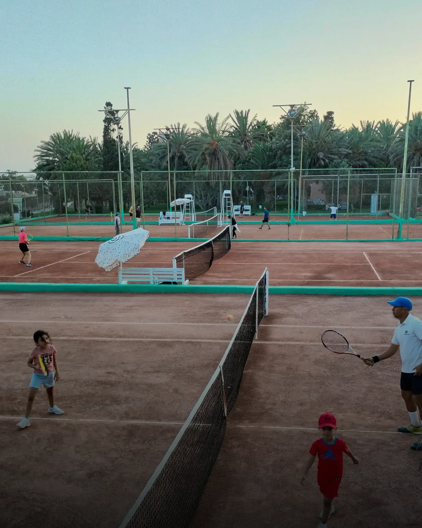 École de tennis jeunes : enfants en séance avec leur coach, filet au premier plan, en fin de journée
