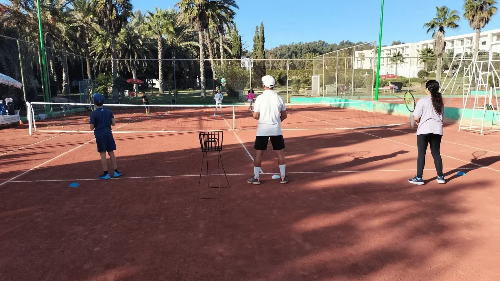 Cours sur la terre battue du club : balles et panier d'entraînement, avec le coach vu de dos face aux joueurs