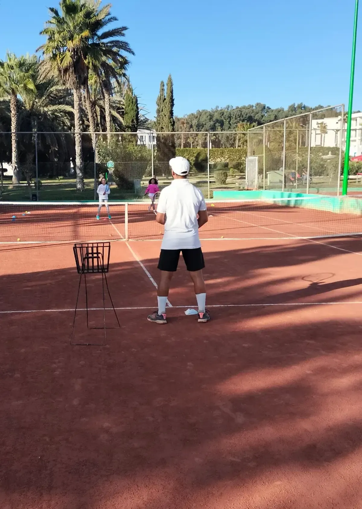 Cours de tennis au club d'Agadir : un coach au panier de balles, ses élèves de l'autre côté du filet, en plein soleil