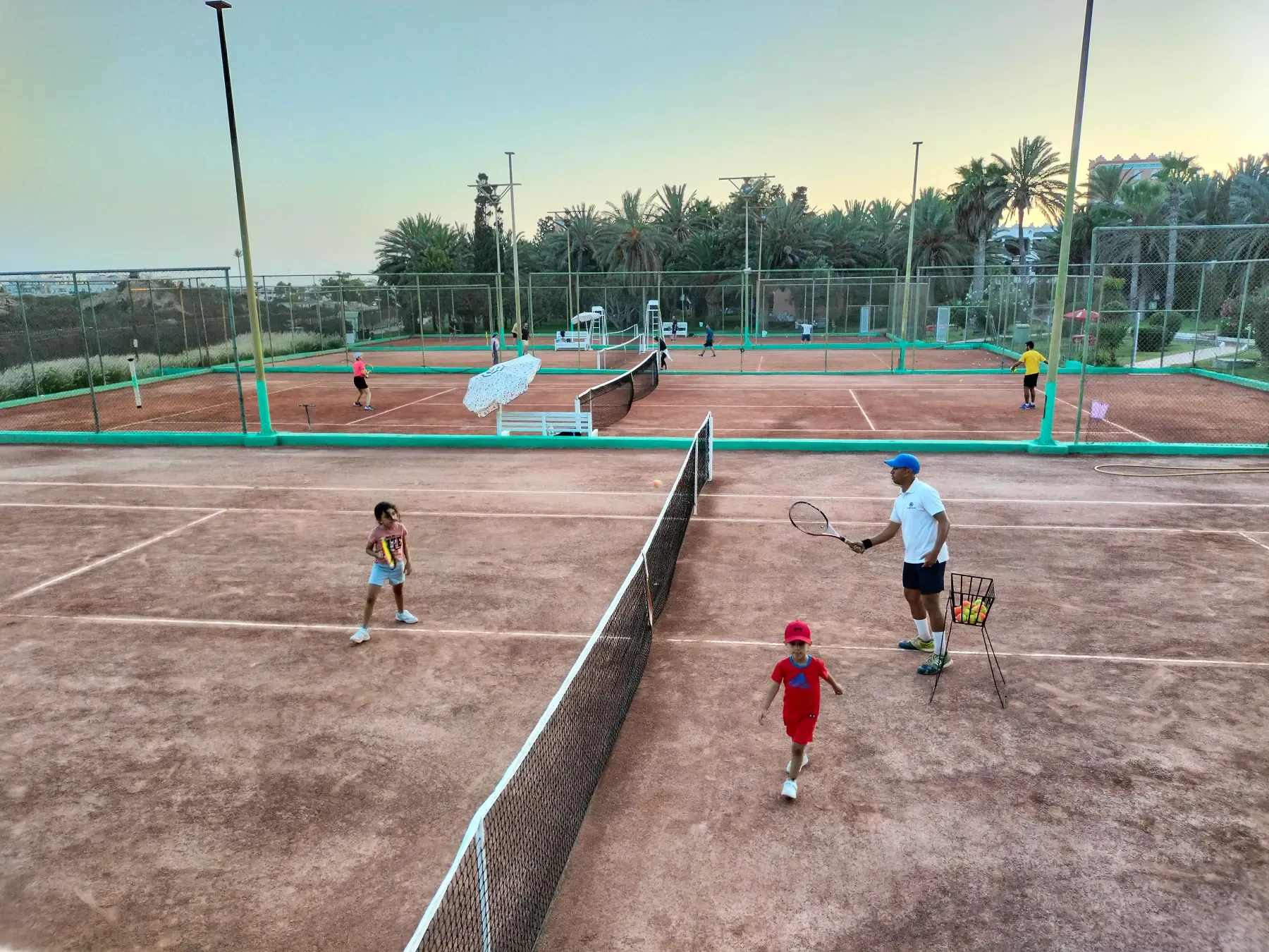 Le club de tennis au cœur d'Agadir : ses quatre terrains en terre battue au crépuscule, un coach et deux enfants au filet
