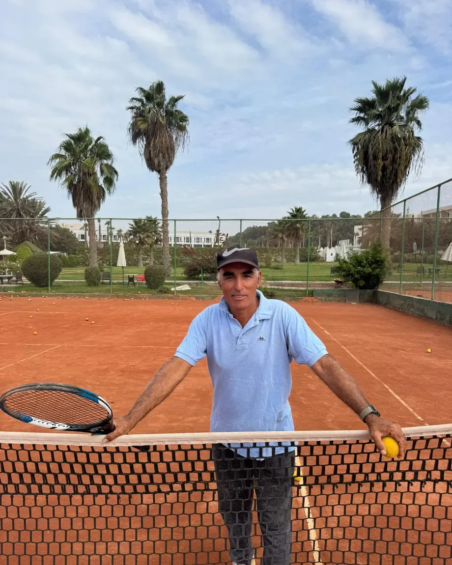 Le coach Hassan, raquette et balle en main, appuyé au filet sur un court du club
