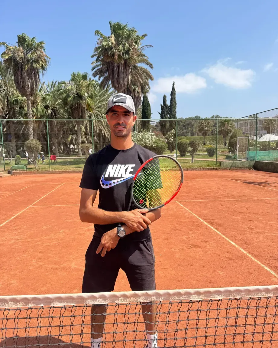 Le coach Imad, casquette et raquette, au filet sur un court en terre battue du club